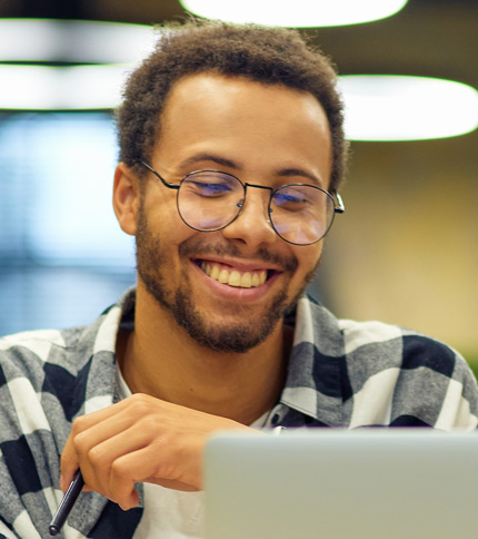 A smiling man with glasses and a short beard sits at a desk, holding a pen and viewing an Accessibility Statement on his laptop. He is wearing a black-and-white plaid shirt and appears to be in a bright, indoor setting.