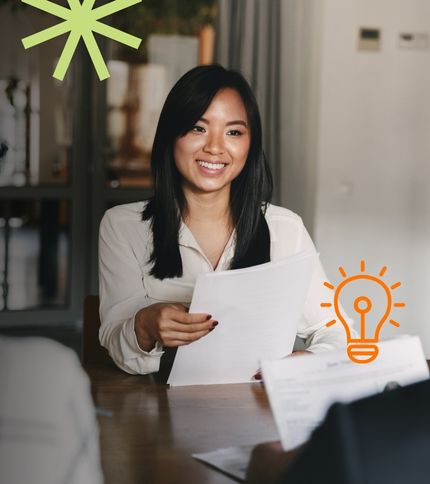 A woman with long dark hair, wearing a white blouse, sits at a table holding papers and smiling during an interview, ready to ace the interview. Decorative graphics of a starburst and lightbulb are overlaid on the image.