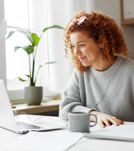 A woman with curly red hair, wearing a grey sweater and glasses on her head, sits at a desk with a laptop, papers about certification, and a grey mug. A potted plant and bright window are in the background.