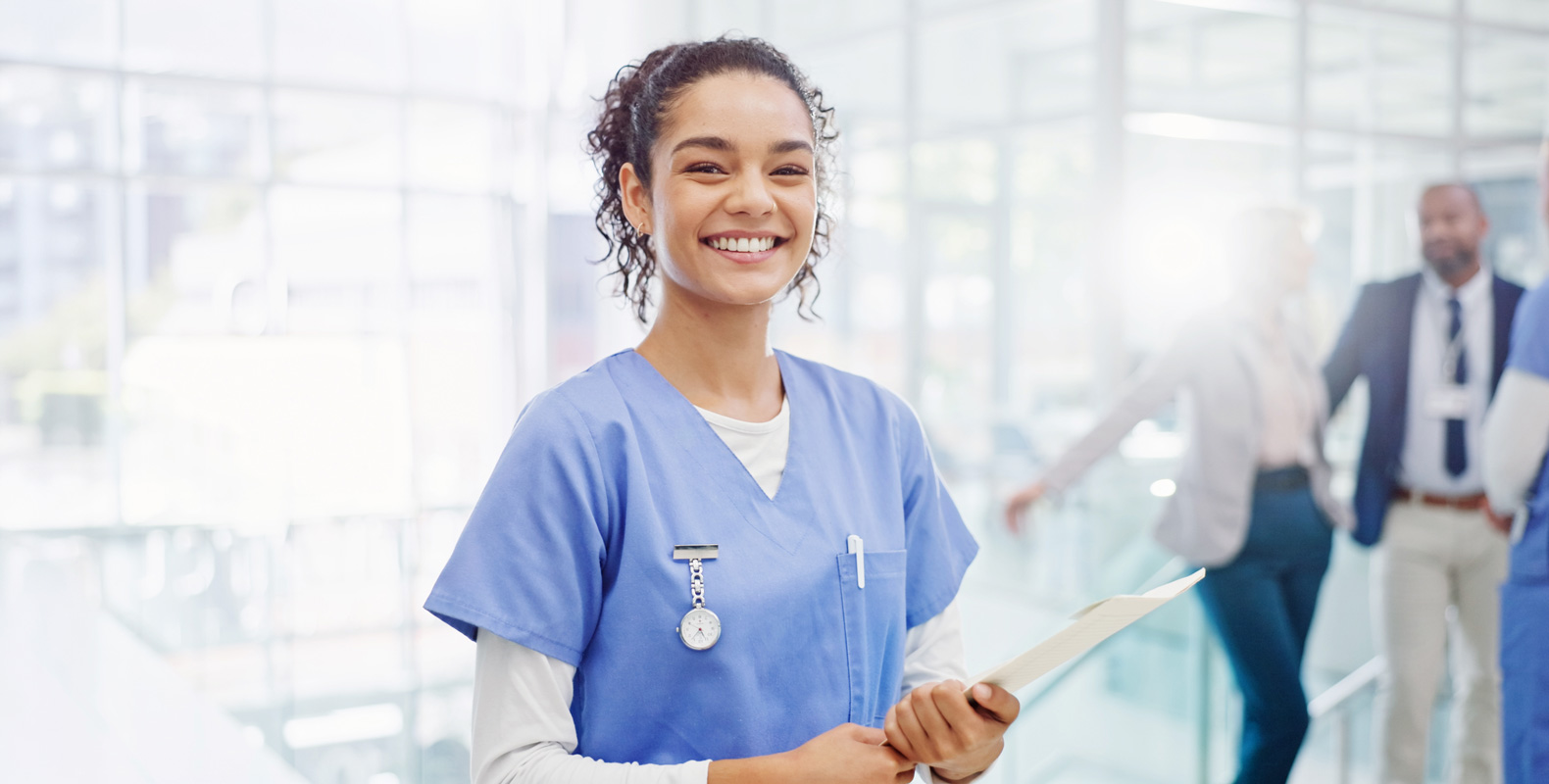 A smiling healthcare worker in blue scrubs holds a clipboard in a bright, modern medical facility, with colleagues conversing in the background.