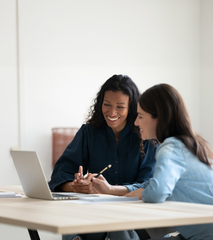 Two women sit at a table, smiling and working together on a laptop. One holds a pencil, and they appear to be discussing something on the screen in a bright, modern office setting.