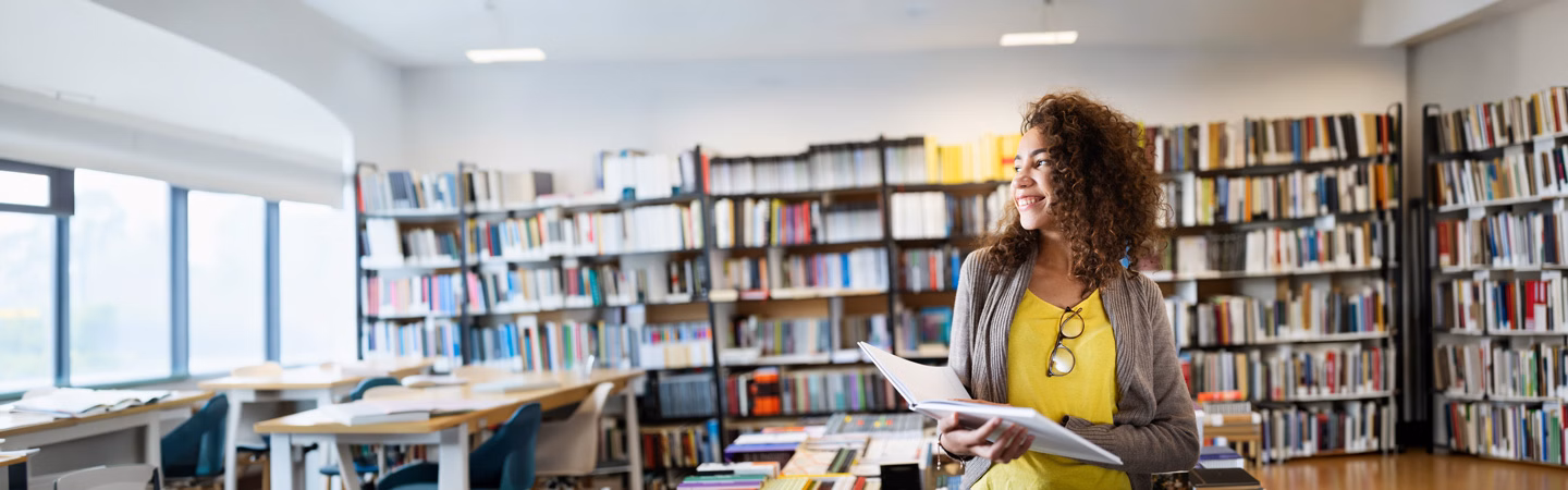 A smiling young woman holding a book in a college classroom library in front of shelves full of books.