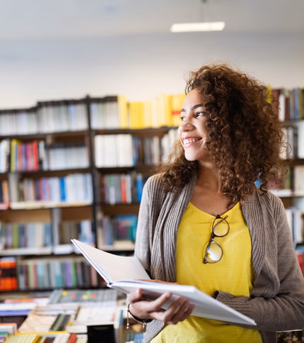 A smiling young woman holding a book in a college classroom library in front of shelves full of books.