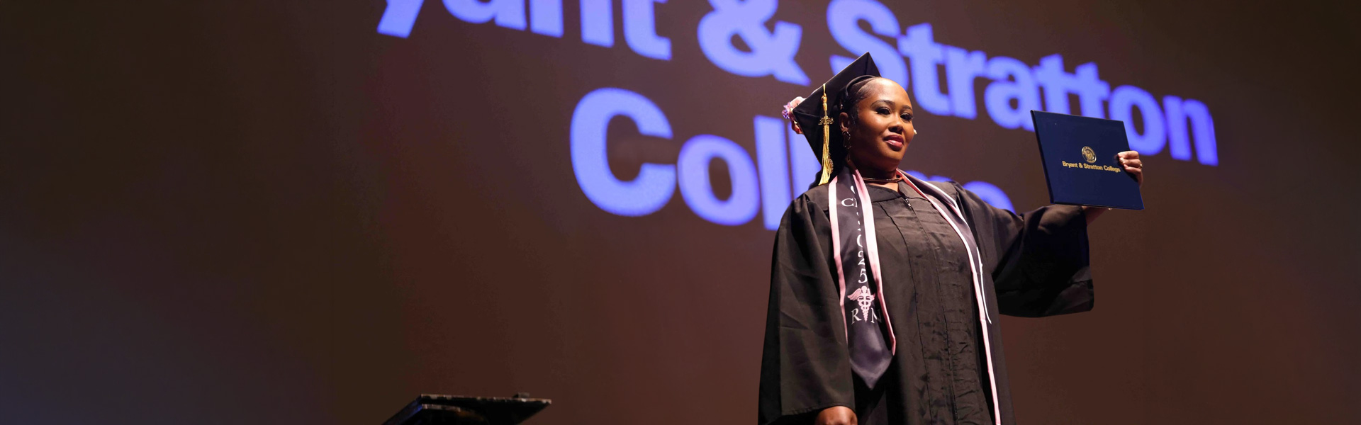 A graduate in cap and gown holds up her diploma on stage, with "Bryant & Stratton College" projected on the wall behind her.