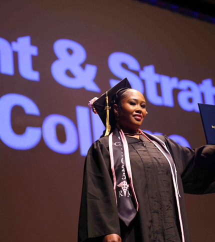 A graduate in a cap and gown proudly holds up her diploma on stage, with large, partially visible text projected on a screen behind her.