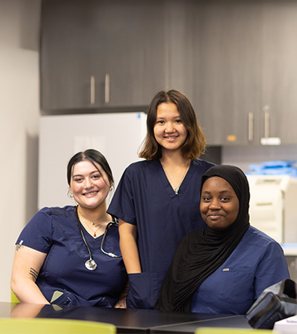 Three women in navy blue medical scrubs, representing healthcare professionals, sit and stand together in a clinical setting, smiling at the camera. One wears a stethoscope and another a black headscarf. Cabinets and equipment are visible in the background.