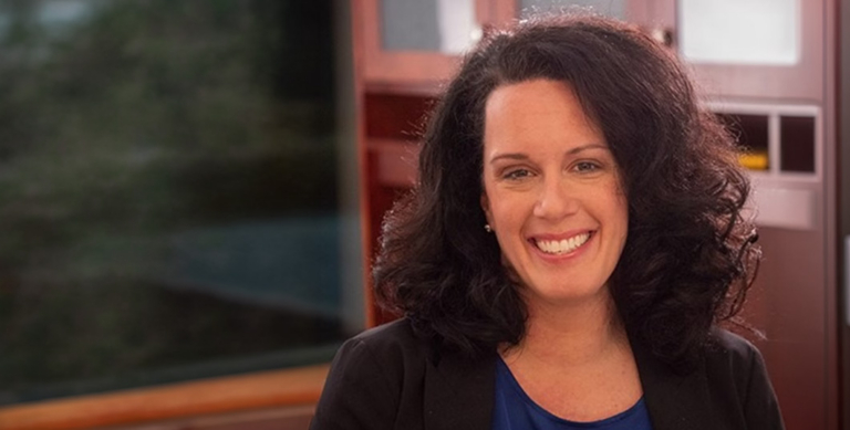 Dr. Kristi Perillo-Okeke, listed in Marquis Who's Who, smiles indoors. She has curly dark hair and wears a dark blazer over a blue top, with a window and wooden cabinets visible in the blurred background.