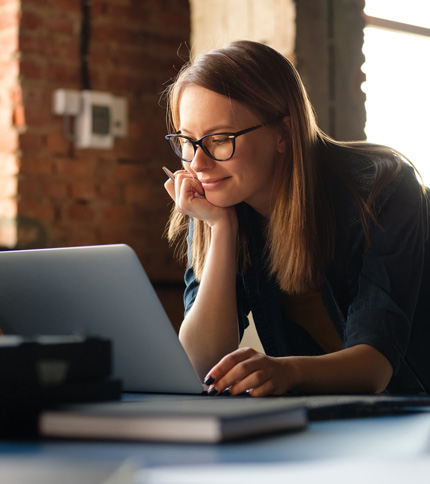 A young woman with glasses is smiling and looking at a laptop screen while resting her chin on her hand. She is sitting at a desk in a room with exposed brick walls and natural light, browsing Admissions FAQs online.