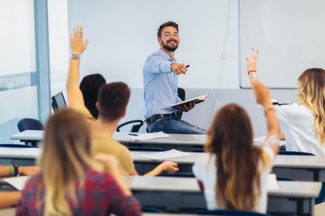 A smiling male teacher sits on a desk at the front of a classroom, holding a book and pointing to students with raised hands, eager to participate in a discussion about Admissions FAQs.
