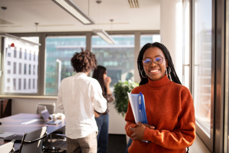 A woman in a rust-colored sweater and glasses smiles while holding folders in a bright office. In the background, two colleagues stand and talk by the window.