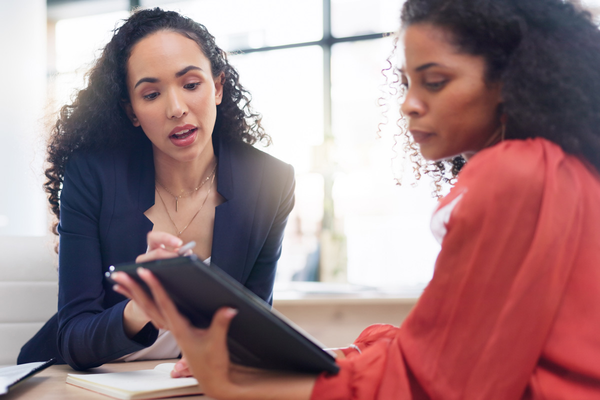 Two women sit together in a bright office space, one in a dark blazer attentively explaining something about the student experience while pointing at a document on a clipboard held by the other, who is wearing a red blouse.