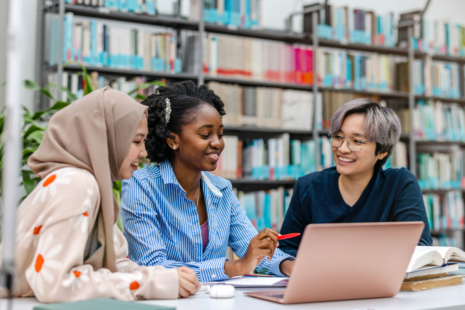 Three students sit together at a table in a library, smiling and working on a laptop as they review Financial Aid Terminology. Bookshelves are visible in the background, and open notebooks filled with Financial Aid FAQs are on the table.