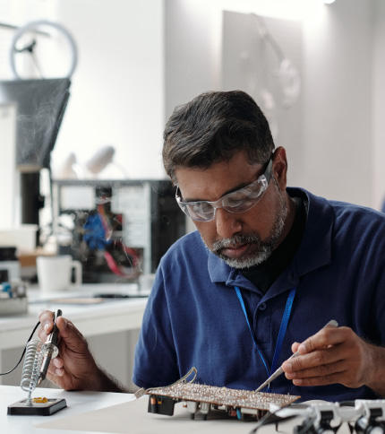 A man wearing safety glasses solders electronic components on a circuit board at a workstation, concentrating on his work in a well-lit lab during an Electronic Technology Experience Day.