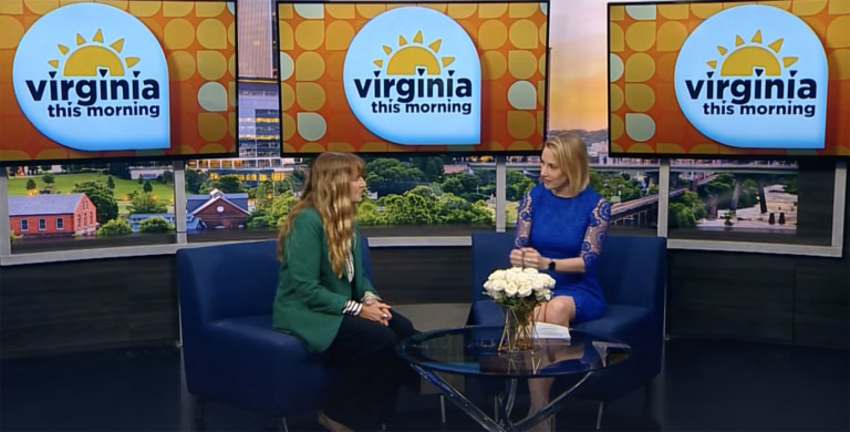 Two women sit on the "Virginia This Morning" TV studio set, discussing Bryant & Stratton College’s Rapid Registration for the Spring Semester. They’re seated in blue chairs with a small table and white flowers between them, three screens showing the show’s logo behind.