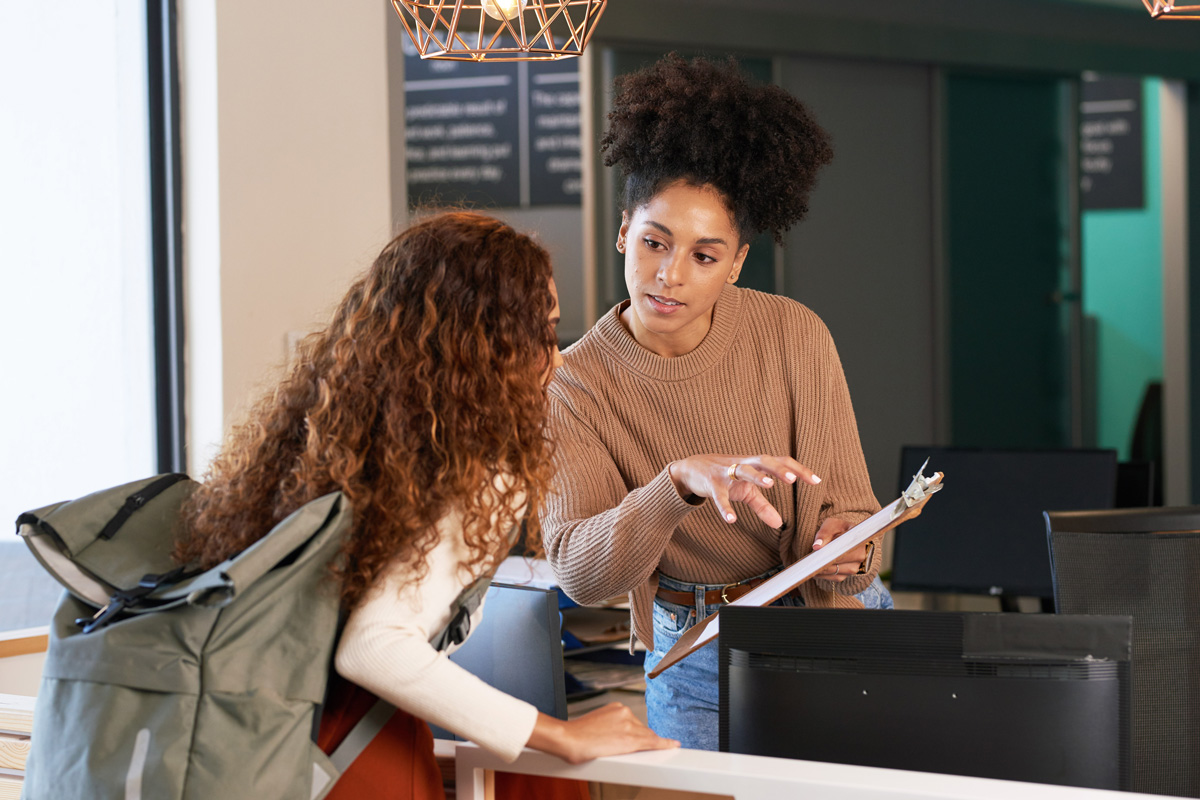 Two women talk in an office setting; one stands holding a clipboard and pointing, while the other, likely seeking guidance on her student experience, leans forward attentively across a counter with her backpack.