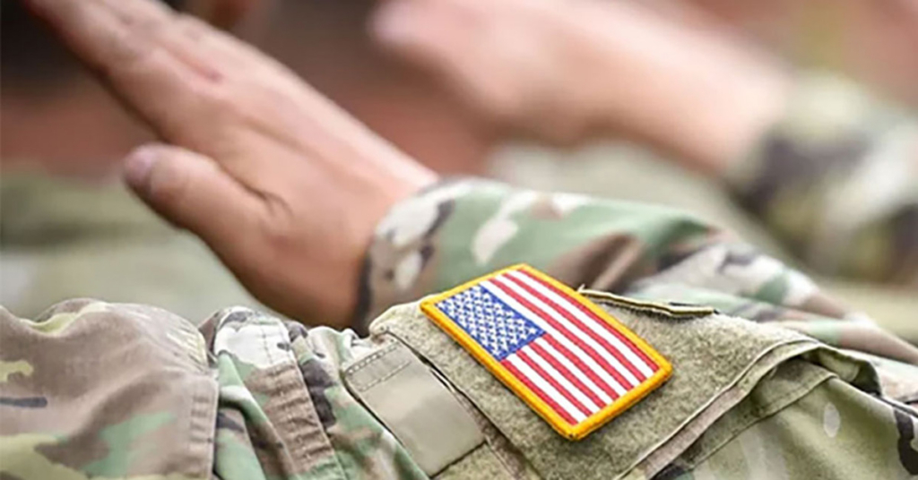 A person in a camouflage military uniform with a U.S. flag patch on the sleeve raises their hand in salute, symbolizing respect and commitment to Military Aid. Another figure in similar attire stands in the background, also saluting.