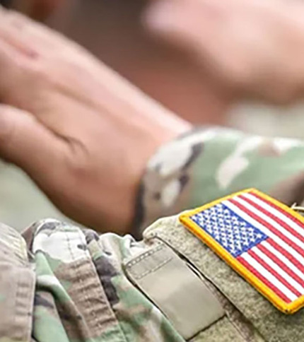 A close-up of a person in military camouflage uniform saluting, with a patch of the American flag visible on their right arm, symbolizing honor and the importance of Military Aid.