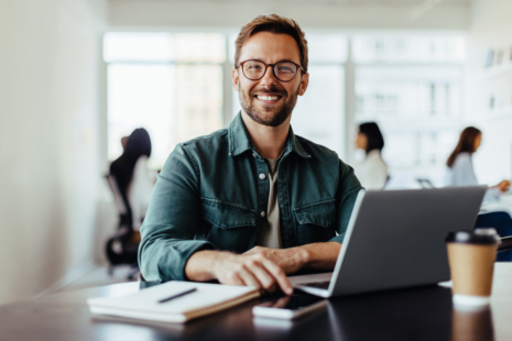 A smiling man with glasses sits at a desk in an office, working on his laptop, researching military aid. A notepad and coffee cup are on the desk, while people work in the background.
