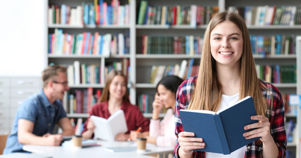 A young woman smiling and holding an open book in a library, possibly researching Private Loans, with three people sitting at a table and reading in the background. Bookshelves filled with books line the walls.