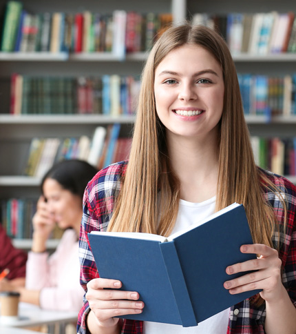 A young woman with long blonde hair smiles and holds an open blue book in a library, perhaps researching Private Loans. Bookshelves are in the background, and another person is studying at a table behind her.