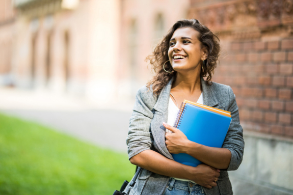 A young woman smiling outdoors, holding colorful notebooks and folders. She stands in front of a brick building, wearing a gray blazer, with curly hair and hoop earrings—perfectly capturing the spirit of returning students at B&SC.