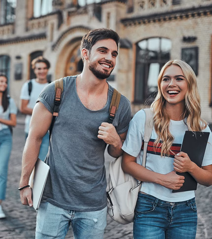 Two smiling college students, one holding a laptop and the other a clipboard, walk together outside a brick building—typical of Returning B&SC Students—while other students are seen in the background.