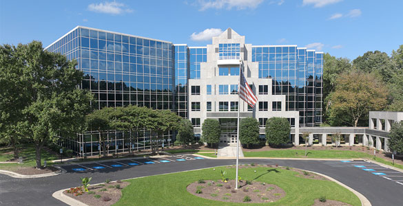 A modern office building with mirrored glass windows and a landscaped entrance suggests a hub for professional development, featuring an American flag, handicap parking spaces, and trees under a blue sky with scattered clouds.