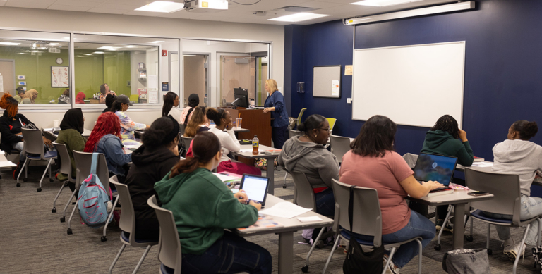 A classroom full of students sits at desks facing a teacher who is standing at the front near a whiteboard. Most students are taking notes or using laptops. The room has bright lighting and large windows.