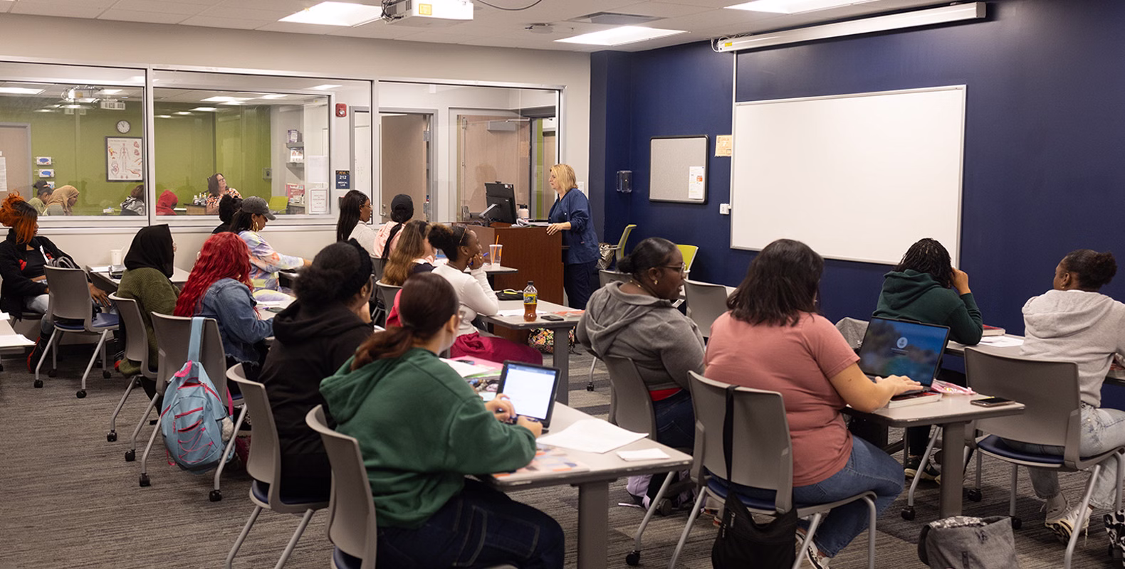 A classroom full of students sits at desks facing a teacher who is standing at the front near a whiteboard. Most students are taking notes or using laptops. The room has bright lighting and large windows.