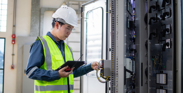 An engineer in a white hard hat and yellow safety vest uses a tablet while inspecting electrical equipment and wiring inside a control panel in an industrial setting.