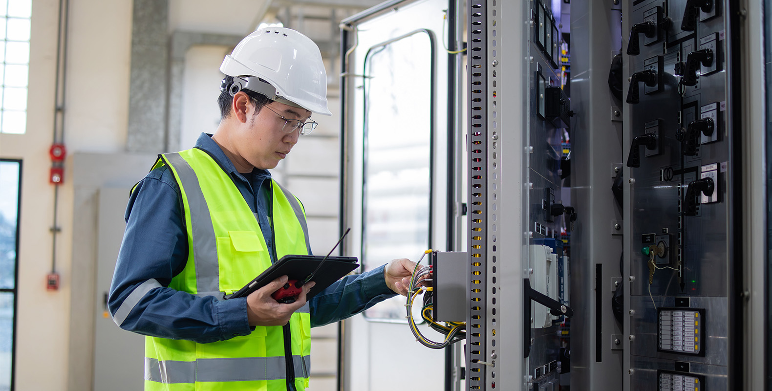 An engineer in a white hard hat and yellow safety vest uses a tablet while inspecting electrical equipment and wiring inside a control panel in an industrial setting.