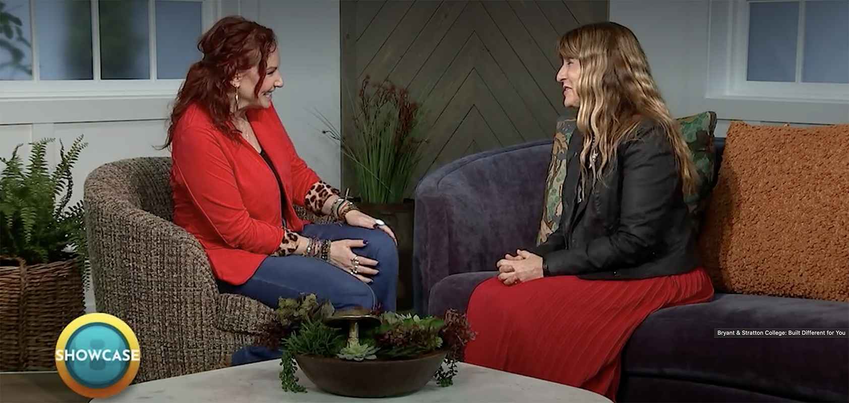 Two women sit facing each other on couches in a cozy studio, engaged in conversation about Rapid Registration at Bryant & Stratton College. One wears a red jacket and jeans, the other a black jacket and red skirt. A "SHOWCASE" logo appears in the bottom left corner.