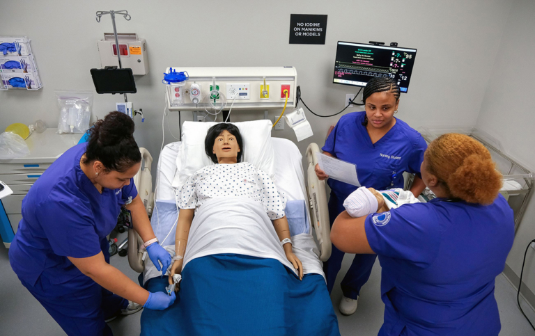 Three nurses in blue scrubs practice medical procedures on a realistic hospital mannequin in a simulated patient room, with medical equipment and monitors in the background—demonstrating the hands-on training students receive as they pursue stackable degrees at Bryant & Stratton College.