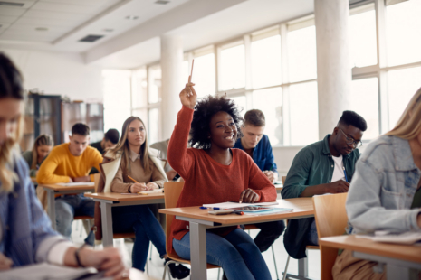 A student raises her hand in a bright classroom, seated among peers pursuing HR degrees or Business degrees. The atmosphere suggests active participation and engagement in learning.