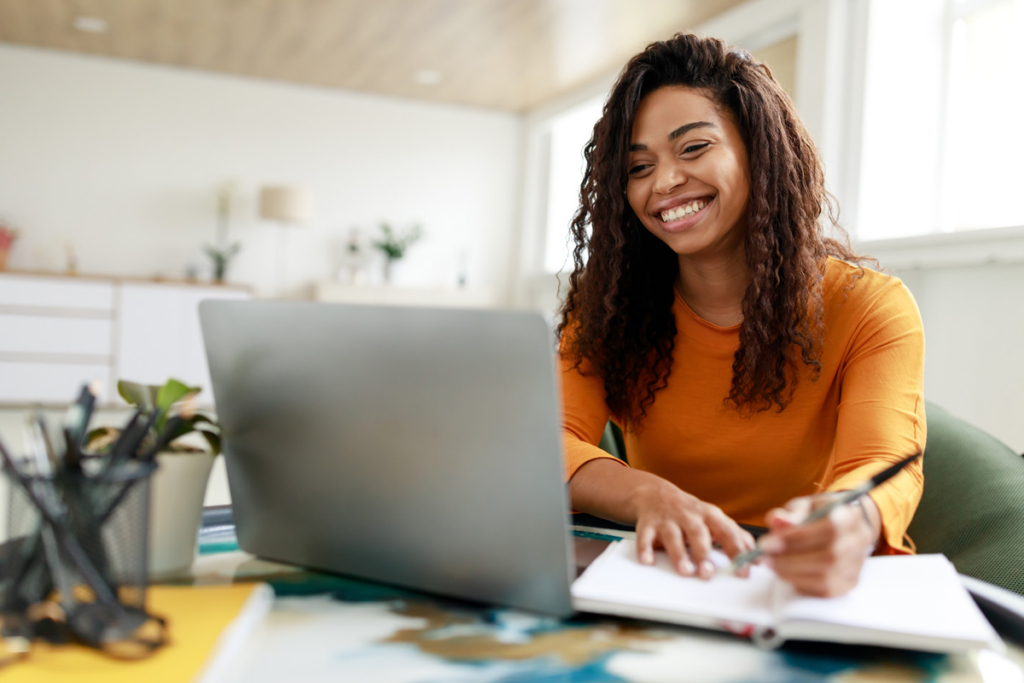 A woman with curly hair, wearing an orange shirt, smiles while working at a laptop and writing in a notebook—perhaps studying for her business degrees—in a bright home office.