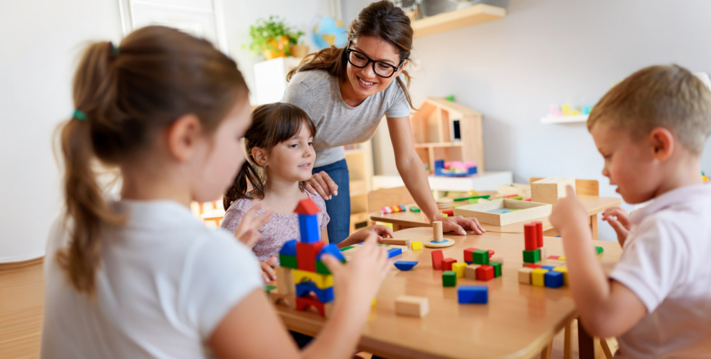 A woman with a CDA Credential smiles as she supervises three young children playing with colorful wooden blocks at a table in a bright, cheerful early childhood education classroom.