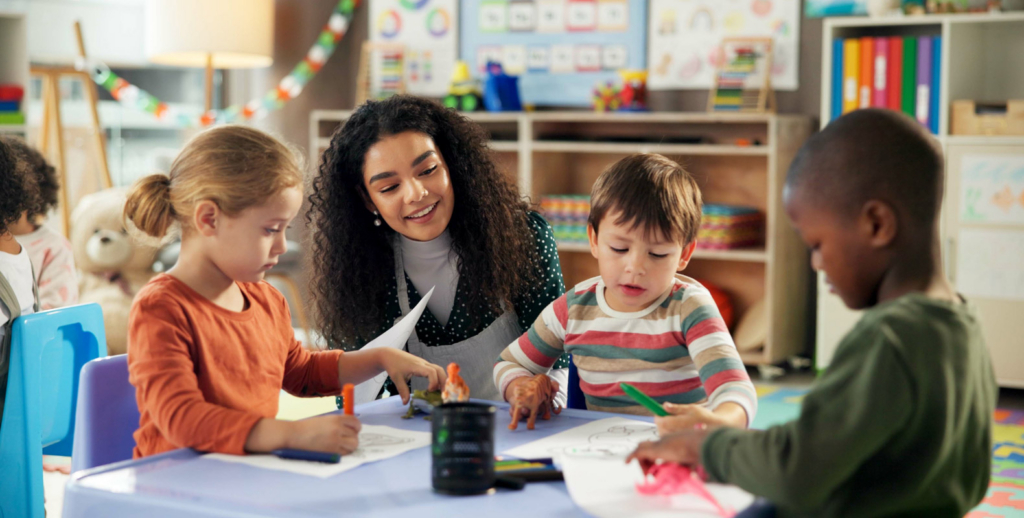 A teacher with a CDA Credential sits at a table smiling and assisting three young children as they draw and color on paper in a colorful early childhood education classroom.