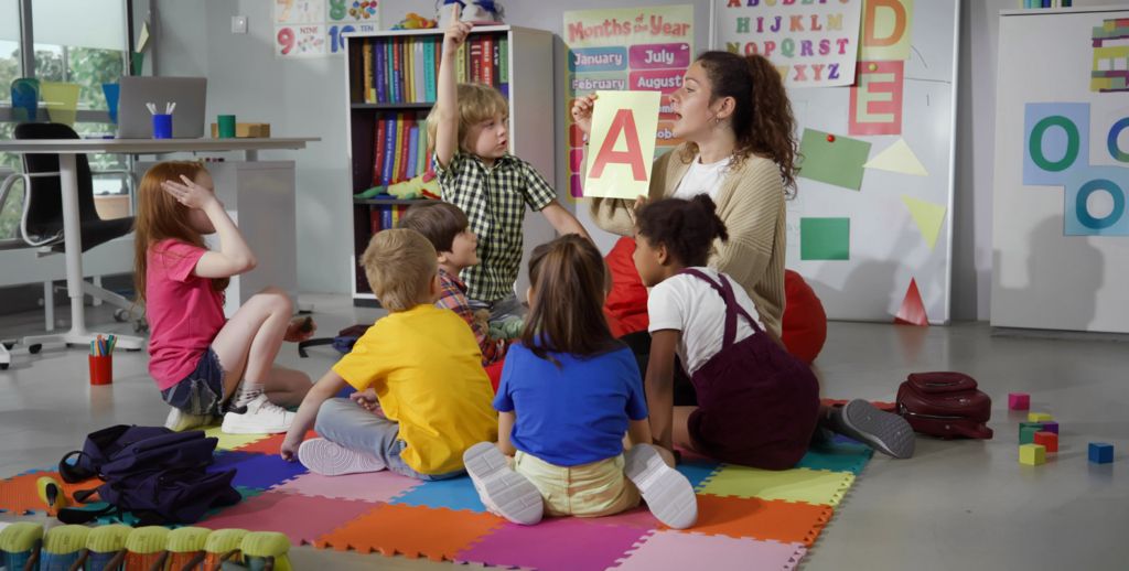 A teacher with a CDA Credential sits on a colorful mat with young children, holding up a card with a large red letter “A.” The children listen attentively, eager to learn in this bright early childhood education classroom setting.