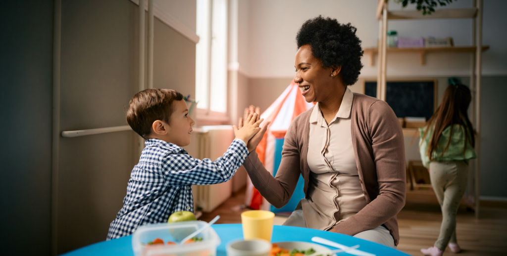 A young boy and an adult woman smile and give each other a high five while sitting at a table in a bright, cheerful room focused on early childhood education, with toys and a play tent in the background. Another child is playing nearby.