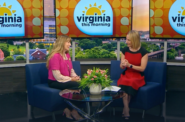Two women sit and talk on the “Virginia This Morning” TV set, discussing Bryant & Stratton College’s Business programs. A small flower arrangement sits on the glass table between them as they share smiles during their conversation.