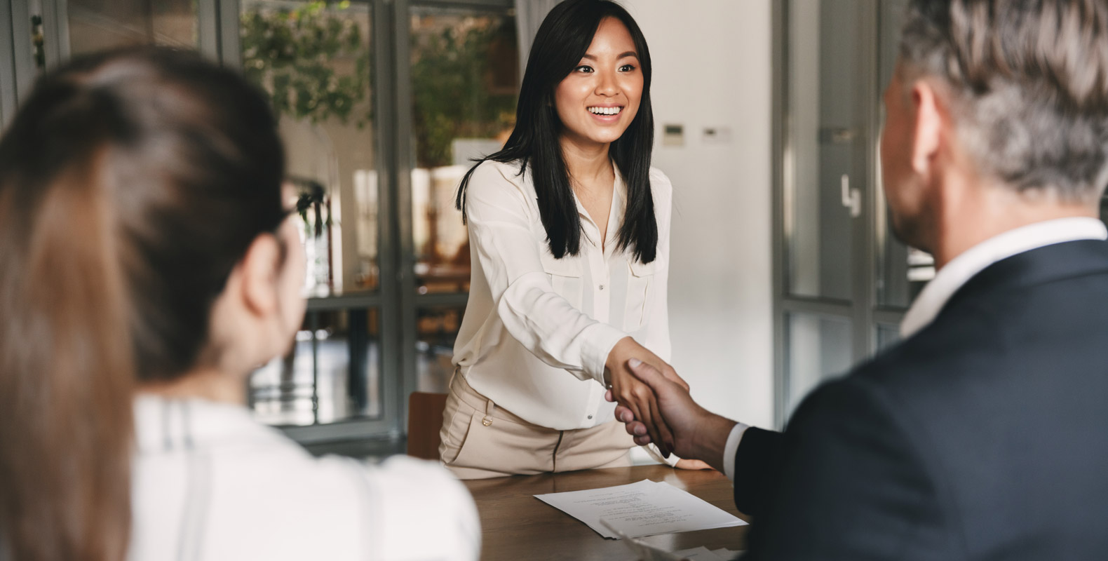 A woman smiles while shaking hands with a man across a table, as another woman looks on. Papers are on the table in a brightly lit office setting.