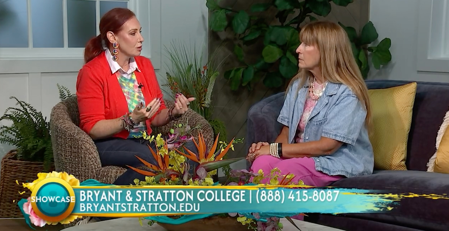 Two women sit in a brightly decorated studio set, engaged in conversation. Text on screen promotes Bryant & Stratton College with a phone number and website, highlighting their partnership with Richmond Region Tourism. Tropical flowers and plants are visible in the foreground.